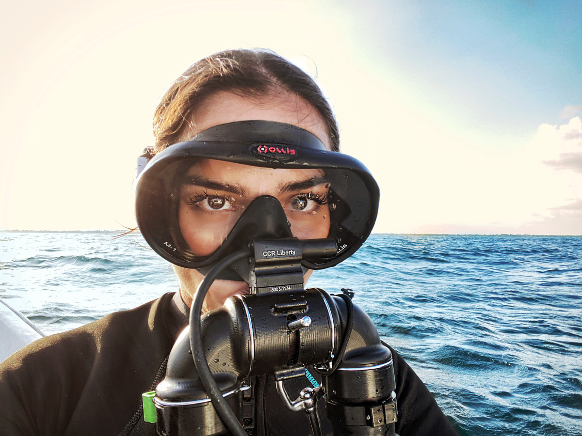 Frau mit schwarzer Tauchermaske und Rebreather-Atemregler vor sonnigem Meereshintergrund.
